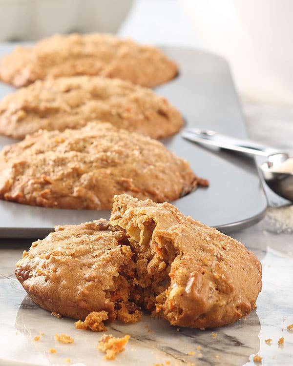 Carrot cake muffin tops on a marble surface, with one broken open to show the crumb, beside a baking pan and measuring spoons.