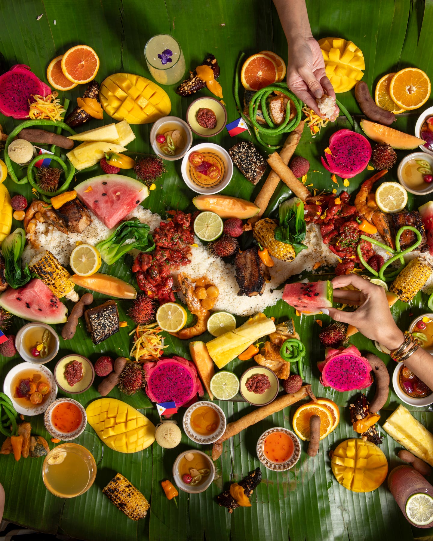 Overhead view of a kamayan-style feast laid on banana leaves, with diners’ hands sharing rice, grilled meats, spring rolls, sauces, and bright tropical fruits like mango, dragon fruit, and watermelon.