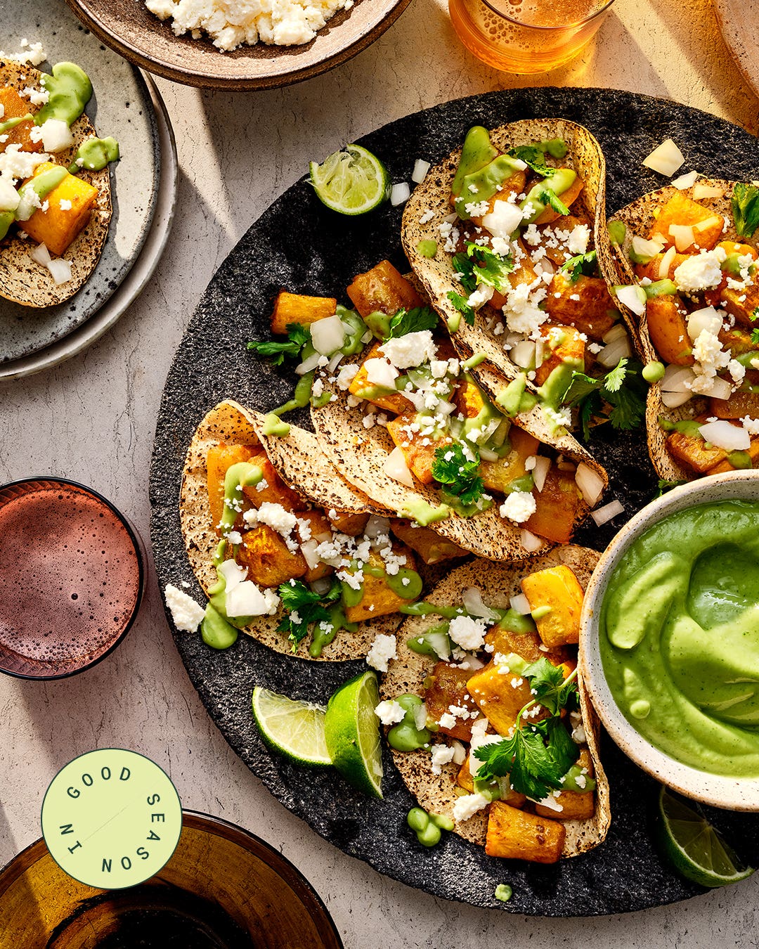 Summer squash tacos on corn tortillas, topped with crumbled queso fresco, diced onion, cilantro, and avocado salsa, with lime wedges and a bowl of green sauce nearby.